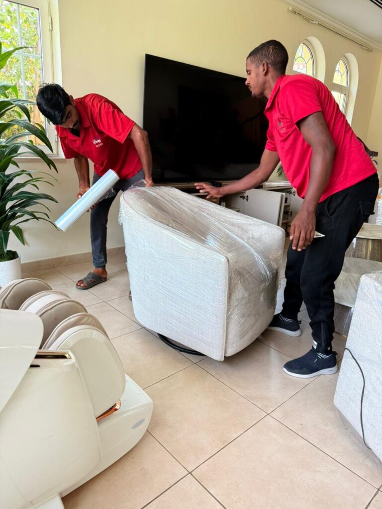 Movers wrapping an armchair in stretch film beside a wall-mounted TV in a Dubai villa